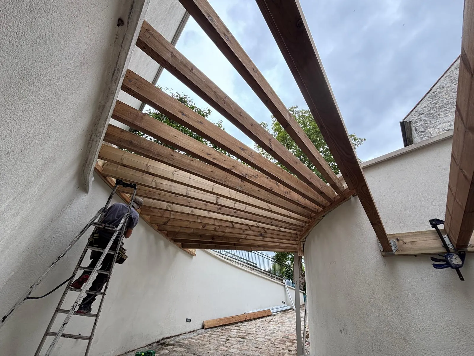Pergola bois en structure ouverte posée en angle au-dessus d'une cour pavée, vue en contre-plongée
