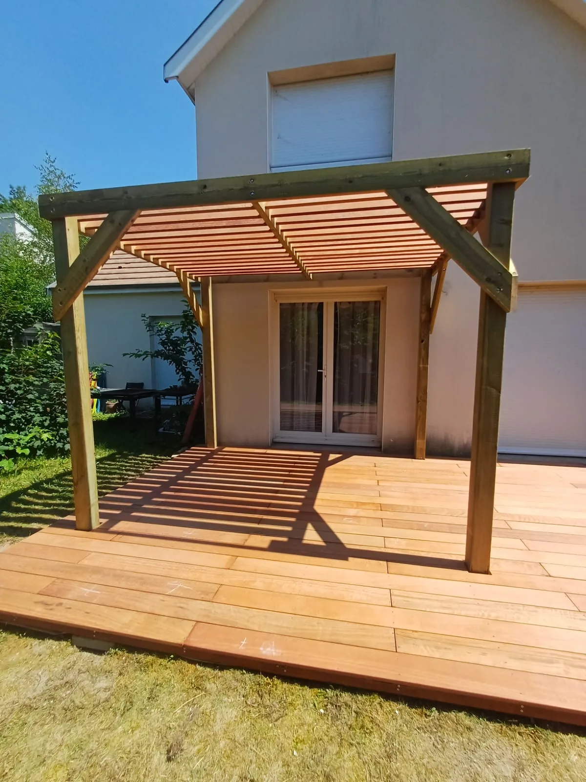 Pergola bois adossée et terrasse en bois exotique bordées de végétation, réalisées par La Planète Bois