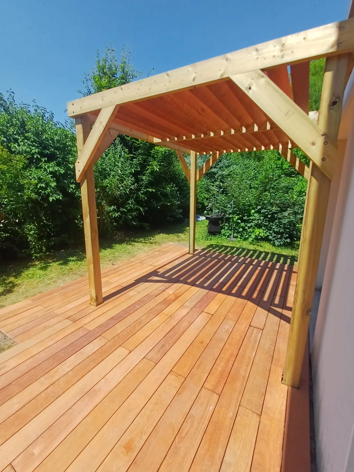 Pergola bois adossée projetant l'ombre de ses chevrons sur une terrasse en bois exotique neuve, réalisée par La Planète Bois